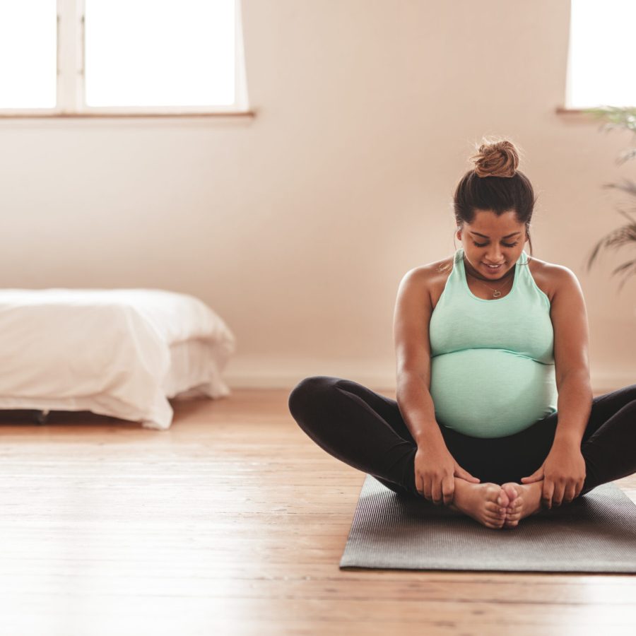 Portrait of young pregnant female working out at home. African pregnant woman practicing yoga at home. Prenatal groins stretch. Buddha Konasana Pose. Pregnancy yoga and fitness.